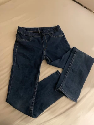 Mörkblå denimleggings med resårmidja - Säljer ett par mörkblå denimleggings med resår i midjan och klassiska jeanssömmar. Byxorna har två bakfickor och är gjorda i stretchigt material som ser ut som jeans men känns som leggings. Perfekta för dig som vill ha en bekväm och snygg look. Barn till tjejer, storlek 122