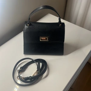 Svart handväska från Gina - Stilren svart handväska i skinnimitation från Gina med guldiga metalldetaljer och avtagbar axelrem. Väskan har en klassisk boxig form, topphandtag och snyggt lås framtill. Perfekt för dig som vill ha en enkel men elegant accessoar. 20x10x17 Finns lite skav på låset