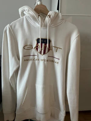 Vit hoodie från GANT med broderad logga - Vit hoodie från GANT med broderad logga och emblem på bröstet. Tröjan har huva med snörning, känguruficka och ribbade muddar. Materialet är mjuk bomull och designen har detaljer i guld och vinrött. Perfekt för en chill och sportig look.
