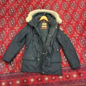 Svart Parajumpers parkas med pälskrage - Säljer en svart Parajumpers kodiak  parkas från Masterpiece Series i storlek XS. Jackan har fluffig pälskantad huva, flera fickor, patch på ärmen och snygga detaljer som dragkedja och knappar. Insidan är quiltad och fodrad för extra värme. Perfekt för kalla dagar.