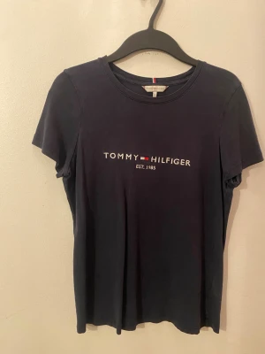 Mörkblå t-shirt Tommy Hilfiger M - Snygg mörkblå t-shirt från Tommy Hilfiger med klassisk logotyp broderad på bröstet. Modellen är kortärmad och har rund hals. Materialet är mjuk bomull som känns skönt mot huden. Perfekt för dig som gillar stilrena och ikoniska plagg.