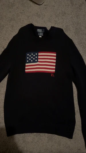 Svart stickad tröja Polo Ralph Lauren USA - Svart stickad tröja från Polo Ralph Lauren med amerikansk flagga i rött, vitt och blått på bröstet samt RL-broderi. Rund hals och långa ärmar, perfekt för dig som gillar klassisk och cool stil.