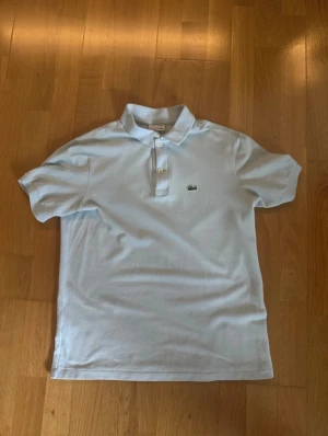 Ljusblå pikétröja från Lacoste - Snygg ljusblå pikétröja från Lacoste med klassisk krage och två knappar framtill. Tröjan har korta ärmar och den ikoniska krokodilloggan broderad på bröstet. Perfekt för dig som gillar stilrena och sportiga plagg.