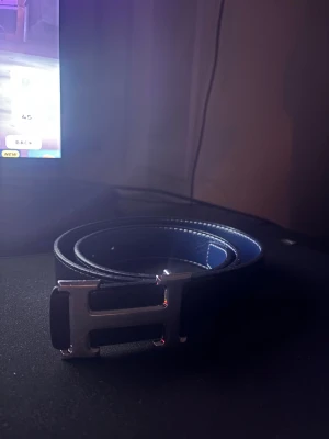 Svart skärp med H-spänne - Snyggt svart bälte med ett stort H-format spänne i silverfärgad metall. Bältet har en stilren design med vita sömmar längs kanterna och är gjort av skinn. Perfekt för att lyfta din outfit med en exklusiv touch.