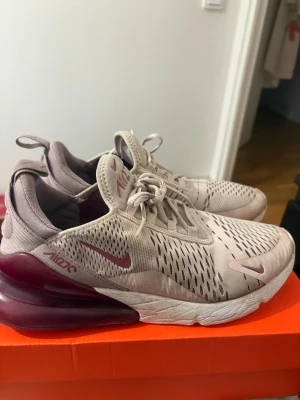 Nike Air Max 270 i beige och vinröd - Nike Air Max 270 sneakers i beige mesh med vinröda detaljer och stor synlig Air-enhet i hälen. Snygg sportig design med snörning och ventilerande ovandel. Perfekta för dig som gillar streetstyle och vill ha bekväma skor med cool look.
