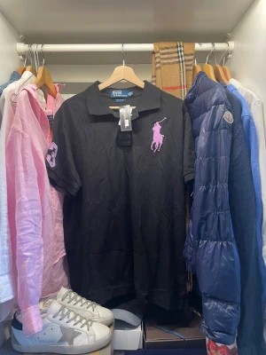 Svart pikétröja Polo Ralph Lauren  - Svart pikétröja från Polo Ralph Lauren i storlek XXL. Tröjan har en stor lila broderad logga på bröstet och siffran 3 på ärmen. Perfekt till en stilren look till vardagen. Nypris ligger runt 1400, tröjan är aldrig använd och har pristagarna kvar.        Skriv vid frågor, priset är inte hugget i sten🤝