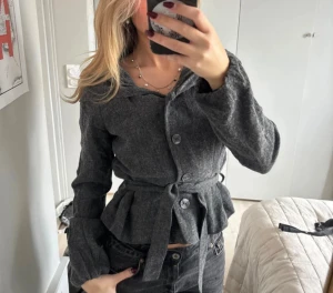 Mörkgrå kofta med bälte - Snygg mörkgrå overshirt med omlottkänsla och knytbälte i midjan. Jackan har stora knappar framtill, långärmade ärmar och ett lätt rynkat midjeparti som ger en cool siluett. Perfekt att styla med jeans för en chill look. Liten i storleken, sitter som en XS!