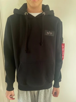 Svart hoodie Alpha Industries - Svart hoodie från Alpha Industries med stor vit logga på ryggen och huvan. Tröjan har snörning vid huvan, ficka framtill och en cool röd tag med texten 'Remove Before Flight' på ärmen. Mjuk bomullskänsla och långärmad passform.