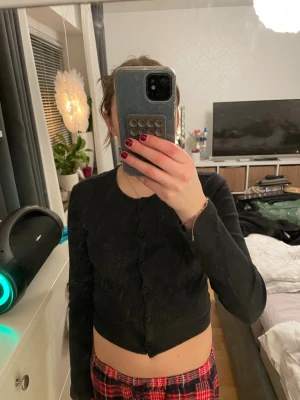 Svart croppad kofta från 157 - Svart ribbad kofta från 157 i croppad modell med långa ärmar och knappar hela vägen framtill. Perfekt att slänga över ett linne eller t-shirt för en chill och snygg look. Enkel och stilren design som funkar året runt.