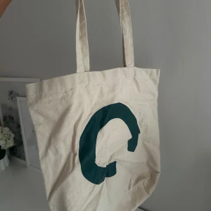 Beige tygkasse med mörkgrönt tryck - Stilren beige tygkasse i bomull med långa handtag och ett stort mörkgrönt C-tryck på ena sidan. Perfekt för att bära böcker, laptop eller shopping. Enkel och trendig design som passar till det mesta.