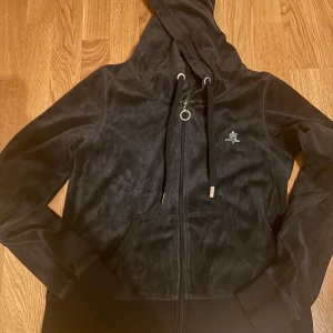 Svart velourhoodie från Cubus med huva - Svart hoodie i mjuk velour från Cubus med dragkedja, huva och snörning. Tröjan har långa ärmar, ribbade muddar och en liten glittrig detalj på bröstet. Perfekt för chill dagar eller när du vill vara extra comfy.