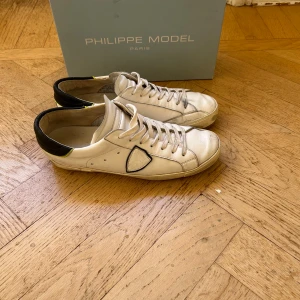 Philippe Model vita sneakers - Snygga vita sneakers från Philippe Model i skinn med svarta detaljer på hälen och en tunn gul linje. Klassisk låg modell med snörning och rund tå. Skorna har märkets ikoniska sköld på sidan och en platt sula för en clean look.