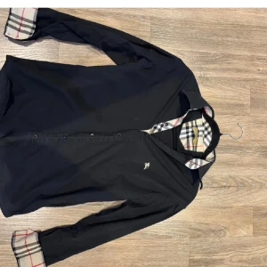 Svart skjorta från Burberry London - (PASSAR XS) Svart skjorta från Burberry London med klassiskt rutigt mönster på insidan av krage och manschetter. Skjortan har långa ärmar, knappar framtill och en liten broderad logga på bröstet. Perfekt för dig som gillar stilrena och tidlösa plagg.