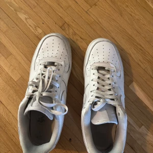 Nike Air Force 1 vita sneakers - Klassiska Nike Air Force 1 sneakers i helvitt skinn med perforerad tå och vit sula. Snygg och tidlös modell med snörning och låg profil, perfekt för dig som gillar streetstyle. Ikonisk Swoosh-logga på sidorna och AIR-detalj på sulan.