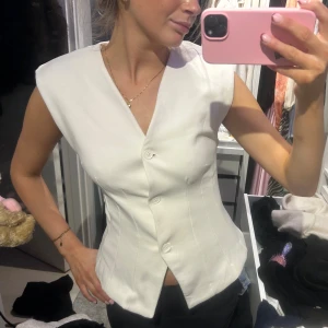 Vit korsettliknande topp med knappar - Säljer en vit korsettinspirerad topp med v-ringning och tre knappar framtill. Toppen är ärmlös och har en figurnära passform med markerade sömmar som ger en snygg siluett. Perfekt för dig som gillar stilrena och trendiga plagg.