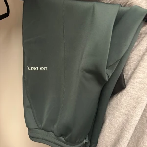 Mörkgröna byxor från Les Deux - Mörkgröna byxor från Les Deux med smal passform och snygg snörning i midjan. Byxorna har raka ben, resår i midjan och diskret logga på ena sidan. Materialet känns mjukt och stretchigt, perfekt för en chill och stilren look. Snöret där man knyter har gått sönder längst ut men annars är de i bra skick
