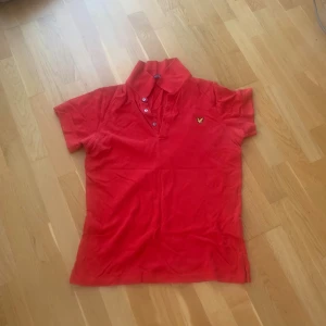 Röd pikétröja från Lyle & Scott - Säljer en klassisk röd pikétröja från Lyle & Scott med gul broderad logga på bröstet. Tröjan har korta ärmar, krage och knäppning med tre knappar. Perfekt för en stilren och sportig look.