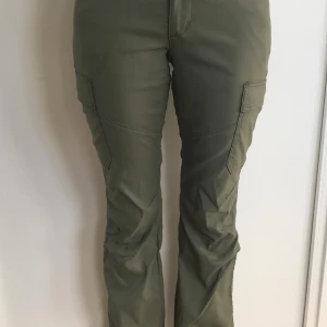 Cargo bootcut byxor Wrangler Stl 26 - Khakigröna cargo/vandrings byxor Wrangler bootcut med fickor på benen och bakfickor. Många fickor bla en liten med dragkedja vid vänster sidficka. Byxorna har en knapp och dragkedja fram. Polyamid/elastan. Stretchiga o sköna.  Använda en gång är i nyskick.  Nypris: 699:-