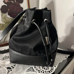 Svart bucket bag med snörning - Snygg svart bucket bag med axelrem och coola metalldetaljer. Väskan har snörning upptill och kombinerar textil med skinnimitation i botten. Perfekt för att ha me i skolan och i klassrummet för att få me småsaker