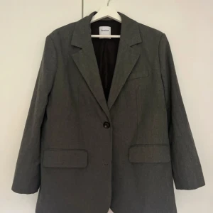 Grå kavaj från Bershka XS - Snygg grå kavaj från Bershka i klassisk modell med en knapp framtill och två fickor med lock. Rak passform och stilren design, perfekt för dig som gillar minimalistiskt och cleant. Fodrad insida och skön att bära. Passar dig som vill ha en enkel men trendig look.