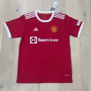 Manchester United röd matchtröja Adidas - Säljer en officiell Manchester United fotbollströja från Adidas i rött med vita detaljer. Tröjan har korta ärmar, rund hals och klubbmärke på bröstet. Materialet är lätt och andas, perfekt för träning eller match. TeamViewer-logga på framsidan.