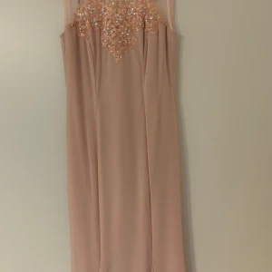 Ljusrosa långklänning med spetsdetaljer - Supersnygg ljusrosa långklänning med transparent mesh upptill och broderade spetsdetaljer med paljetter över bröstet. Klänningen är ärmlös och har en rak, elegant siluett med dragkedja i ryggen. Perfekt för speciella tillfällen när du vill sticka ut lite extra.