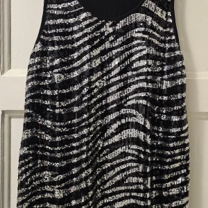 Glittrig svart och silver kortklänning - Snygg kort klänning med glittriga paljetter i svart och silver som skapar ett zebramönster. Ärmlös modell med rund halsringning och mjuk svart baksida. Perfekt för dig som vill sticka ut med en cool och festlig look.