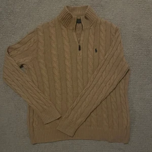 Beige kabelstickad tröja Polo Ralph Lauren - Kabelstickad beige tröja från Polo Ralph Lauren med halv dragkedja och hög krage. Klassisk design med ribbade muddar och det ikoniska broderade logot på bröstet. Perfekt för lager-på-lager under kyliga dagar. Nypris: 2400kr✅💯