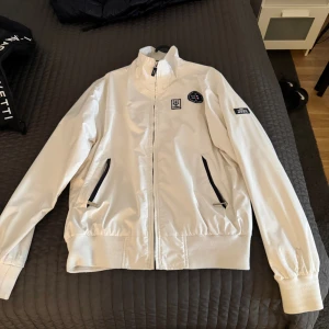 Vit vindjacka från Gant Yacht Club - Vit vindjacka från Gant Yacht Club med coola marina patchar på bröstet och ärmen. Jackan har dragkedja framtill, två fickor med svarta dragkedjor och ribbade muddar. Perfekt för dig som gillar nautisk stil och snygga detaljer.