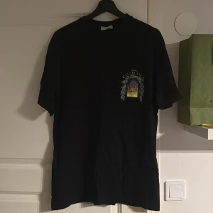 Casablanca tshirt - Svart t-shirt från Casablanca Tennis Club i storlek M. Framsidan har ett litet tryck med texten 'Avenida Casablanca' och ett motiv av en byggnad med blad. Baksidan har ett större tryck med samma motiv och text. T-shirten är i mjuk bomull och har en normal passform.