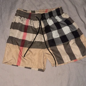 Burberry bad shorts - Snygga shorts med klassiskt rutigt mönster i beige, svart, vitt och rött. De har elastisk midja och svart snörning framtill. Perfekta för sommaren och har en loose passform. Materialet är lätt och syntetiskt, vilket gör dem sköna och luftiga.