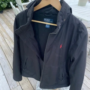 Svart vindjacka från Polo Ralph Lauren - Svart vindjacka från Polo Ralph Lauren med huva och dragkedja framtill. Jackan har två fickor med dragkedja och röd broderad logga på bröstet. Materialet är 100% polyester och insidan har mjuk viskos. Perfekt för dig som gillar stilren och klassisk look. OBS. Dragkedjan är sönder men det går att laga. 