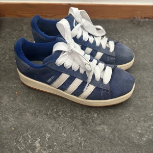 Adidas Campus blå sneakers mocka - Säljer ett par klassiska Adidas Campus sneakers i blå mocka med vita ränder och vit sula. Skorna har rund tå, platt sula och vita snören. Perfekta för dig som gillar retrostil och vill ha något som sticker ut lite extra.