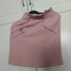 Rosa kortkjol med dragkedjor från NLY Trend - ⚠️ Skriv till mig innan köp!⚠️Säljer en ljusrosa kortkjol från NLY Trend i storlek 34. Kjolen har tre snygga silverfärgade dragkedjor framtill och en diskret dragkedja bak. Stilren och enkel design i ett mjukt syntetmaterial, perfekt för att lyfta din outfit.