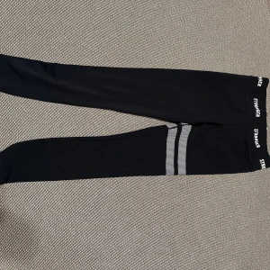 Svarta leggings från Stronger, S - Använda ett fåtal gånger! Svarta leggings från Stronger i storlek S med vita loggor i midjan och två vita ränder på ena benet. Snygg och sportig design, perfekt för gymmet eller löpning. Materialet är stretchigt och följsamt för maximal komfort.
