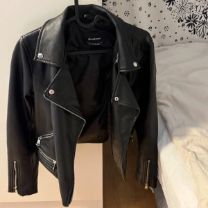 Svart fejkskinnjacka från Stradivarius - Snygg svart skinnjacka från Stradivarius med klassisk bikerstil. Jackan har silvriga dragkedjor på fickor och ärmar, bred krage med tryckknappar och coola detaljer vid midjan. Perfekt för dig som vill ha en edgy look och gillar streetstyle. Obs fejkskinn