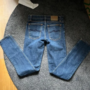 Blå slim jeans från Nudie storlek 28/32 - Säljer ett par klassiska blå skinny/slim jeans från Nudie Jeans med snygga detaljer på bakfickorna och orange sömmar. Jeansen har normal passform och är tillverkade i mjukt bomullsmaterial. Perfekta för dig som gillar en smal siluett och stilren look. Det är 9,5/10 skick, ser helt nya ut nästan😇. Har slim passform och passar dig som är mellan 160-165. Säljer då de är försmå! Skriv vid frågor och funderingar!✨Nypris 1899💸🤝