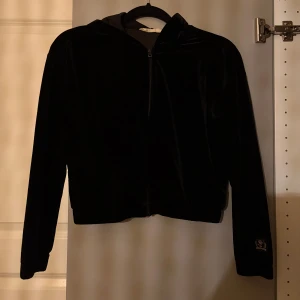 Svart velour hoodie med dragkedja - Svart hoodie i mjuk velour med huva och hel dragkedja framtill. Långärmad modell med broderad logga på ena ärmen. Perfekt för chill dagar och har en skön loose passform.