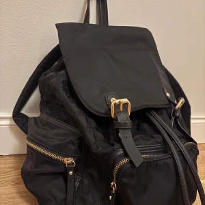 Svart ryggsäck med gulddetaljer - Snygg svart ryggsäck med guldfärgade dragkedjor och spännen. Väskan har flera ytterfack med dragkedja, snörning upptill och lock med spännen. Även ett stängt innerfack och två öppna innerfack. Perfekt för dig som vill ha en praktisk och stilren väska där mycket får plats.