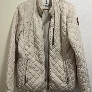 Beige quiltad jacka Iconic Sport L - Snygg beige quiltad jacka från Iconic Sport i storlek L. Jackan har dragkedjor på fickorna och bröstet, ståkrage och ett stilrent rutmönster. Perfekt för dig som gillar en clean och sportig look.