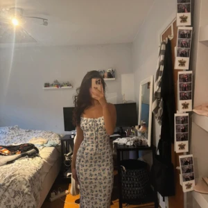 Blommig långklänning med knytband - Superfin långklänning i vitt med blått blommönster och smickrande passform. Klänningen har smala axelband med knyt och en söt snörning framtill vid bysten. Perfekt för sommarens alla tillfällen!