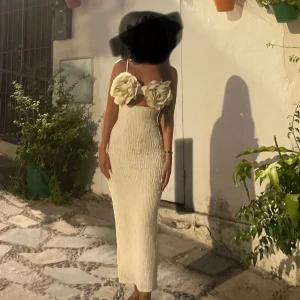 Vit ribbad långklänning med rosdetaljer - Superfin vit långklänning med ribbad struktur och stora rosdetaljer på bysten. Klänningen har smala axelband och en figurnära passform som framhäver siluetten. Perfekt för dig som vill sticka ut med en unik och romantisk look.