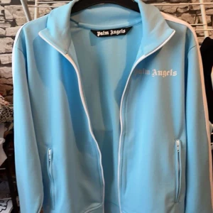 Palm Angels ljusblå hoodie  - Palm Angels ljusblå vindjacka med vit dragkedja och vita ränder längs ärmarna. Jackan har ståkrage, två fickor med dragkedja och logotypen tryckt på bröstet. Snygg sportig look med svart detalj på ärmen.