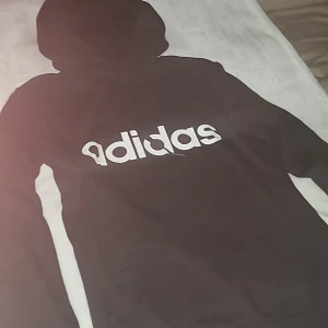 Svart Adidas hoodie - Svart hoodie från Adidas i storlek XS med stor vit Adidas-logga på bröstet och liten logga på ärmen. Klassisk känguruficka framtill och huva. Perfekt för en sportig och chill stil.