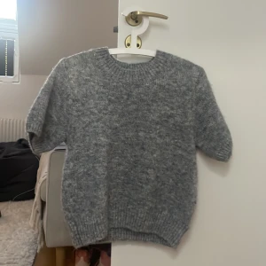 Grå stickad tröja från Gina Tricot - Mysig grå stickad tröja från Gina Tricot med korta ärmar och rund halsringning. Tröjan har en fluffig känsla och ribbade muddar vid ärmslut och nederkant. Perfekt för lager-på-lager under kyliga dagar.