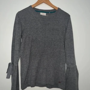 Grå höst tröja med knyt från Holly & White - Mysig grå långärmad tröja från Holly & White med snygga knytband på ärmarna. Tröjan har en enkel rund halsringning och är tillverkad i mjukt material som passar perfekt till höst och vinter. Har aldrig använts. Stilren och lätt att matcha med det mesta. Tröjan material: Bomull och akryl. 