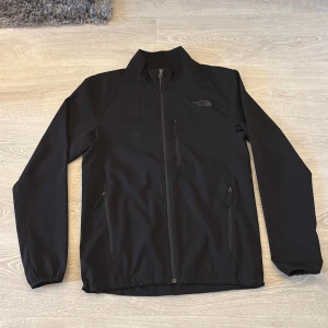 Svart vindjacka från The North Face - Svart vindjacka från The North Face med diskret logga på bröstet. Jackan har hel dragkedja framtill, tre fickor med dragkedja och hög krage. Tillverkad i slitstarkt syntetmaterial och har en clean, sportig look som funkar året runt.
