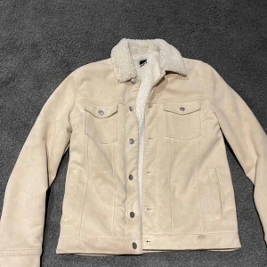 Beige teddyjacka med fuskpäls-krage - Snygg beige teddyjacka från W.BXTR med mjuk fuskpäls på kragen och insidan. Jackan har två bröstfickor med knappar, raka fickor på sidorna och stängs med tryckknappar framtill. Perfekt för kyliga dagar och ger en clean look.