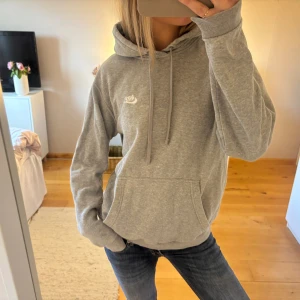 Grå hoodie från Nike med huva - Grå hoodie från Nike med broderad vit logga på bröstet. Tröjan har huva med snörning, stor magficka och långa ärmar. Mjuk och skön bomullsmix som passar perfekt till chill eller streetwear. Klassisk loose passform och stilren look.