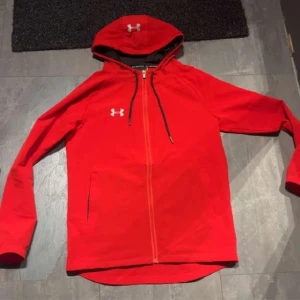 Under armour windbreaker - Riktigt snygg röd vindjacka från Under Armour med huva och hel dragkedja. Jackan har svarta detaljer, två fickor framtill och logga på bröstet och huvan. Perfekt för dig som vill ha en sportig look och gillar streetstyle.